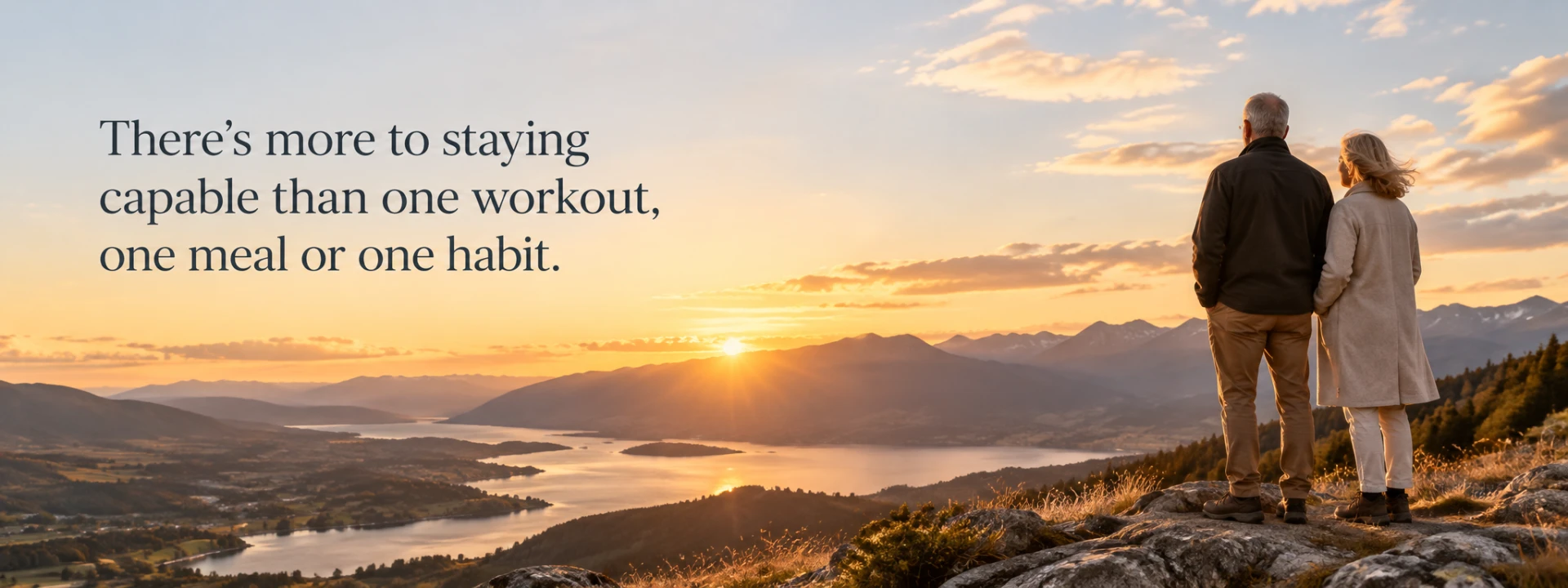 An older couple standing together on a rocky ridge at sunrise, looking over a wide valley of lakes and distant mountains, with the words: There’s more to staying capable than one workout, one meal or one habit.
